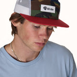 male model wearing a mesh camp hat with the till i die logo woven on the front. The hat has a classic camo from, navy top, red brim, and white mesh sides