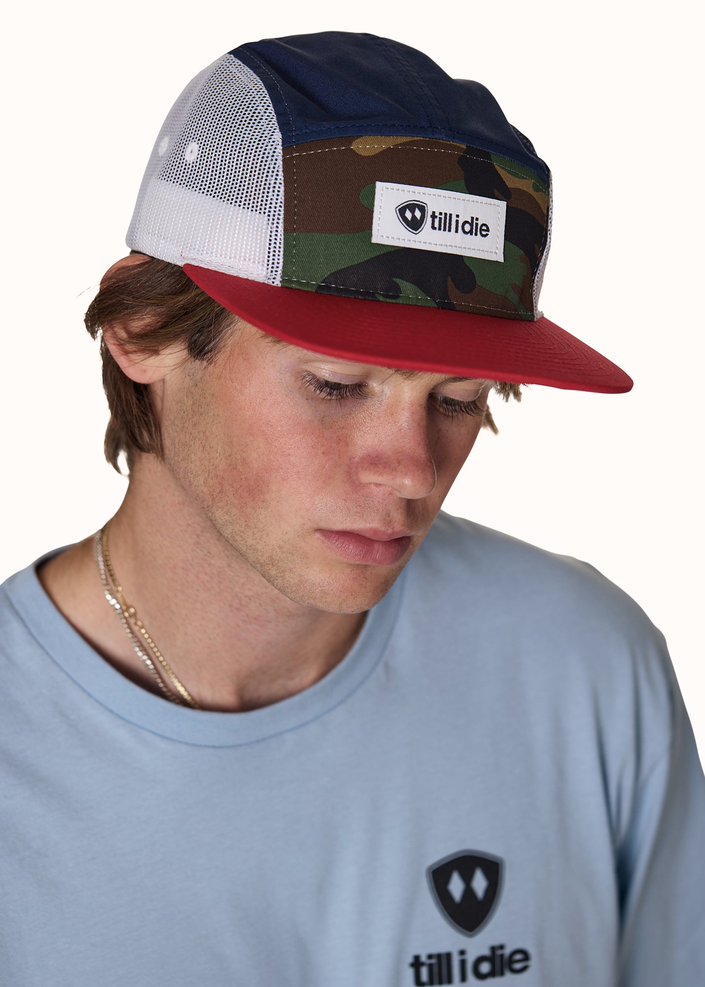 male model wearing a mesh camp hat with the till i die logo woven on the front. The hat has a classic camo from, navy top, red brim, and white mesh sides