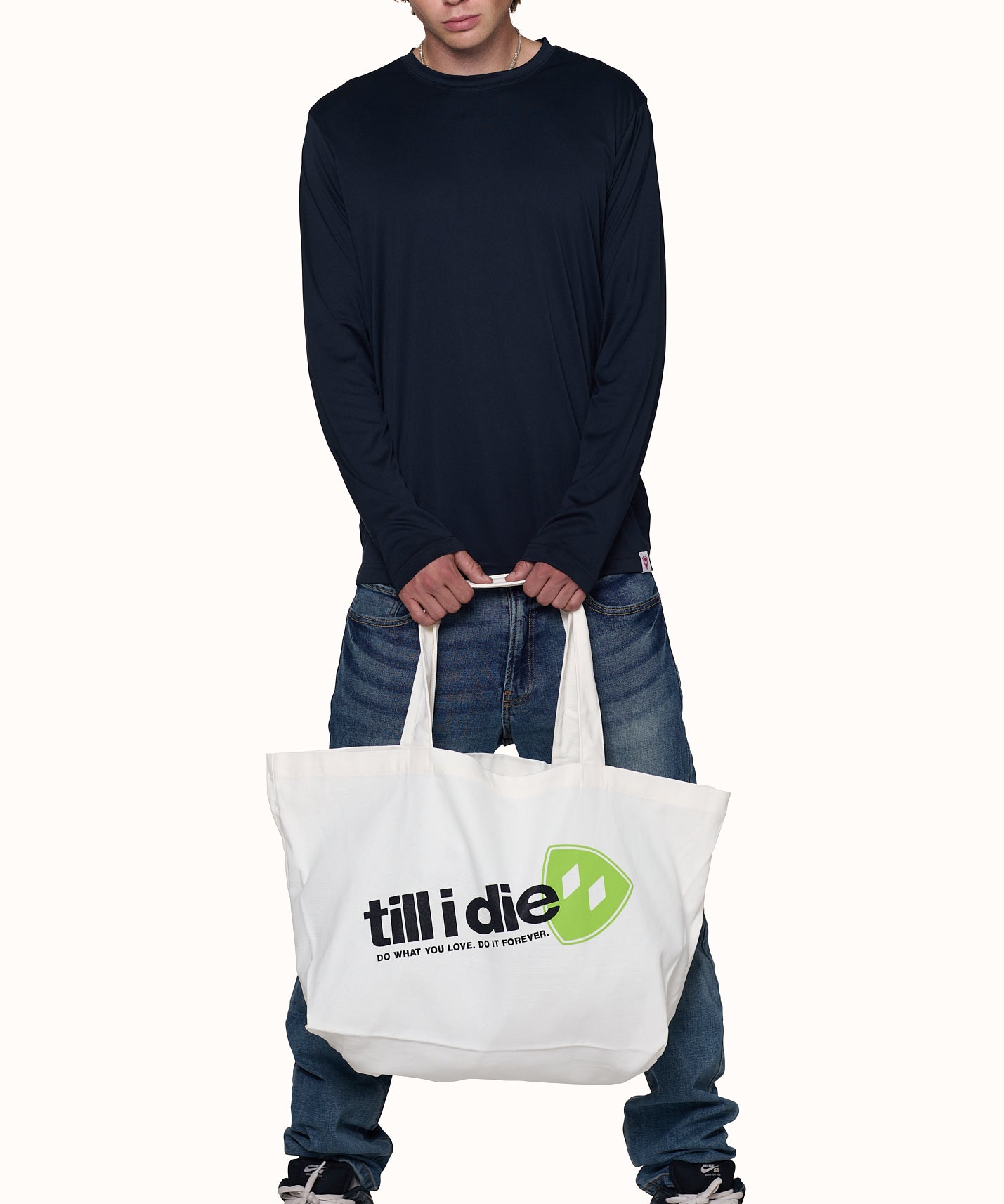 male model wearing a black performance long sleeve holding a natural color canvas tote featuring Till i die text, lime green shield, and "do what you love. do it foever"