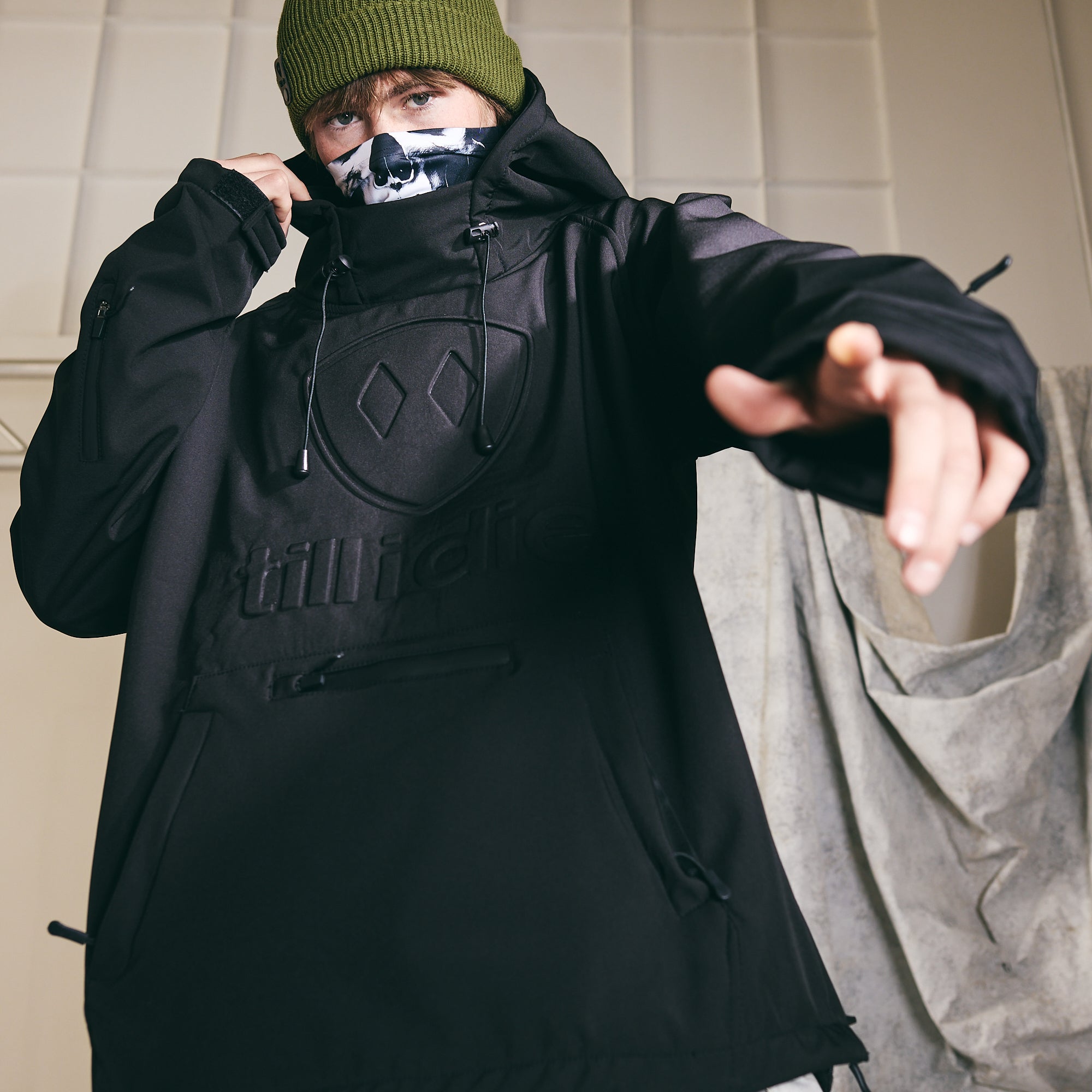 Male model wearing the Skull Neck Gaiter, black Performance Pullover Jacket, and Military Green Knit Beanie. He grips the jacket collar with one hand while pointing directly at the camera. Set against an industrial backdrop, the lighting emphasizes the embossed Till I Die logo on the front of the jacket.