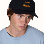 male model wearing a black snapback cap with an embroidered orange skateboard graphic with "till i die"
