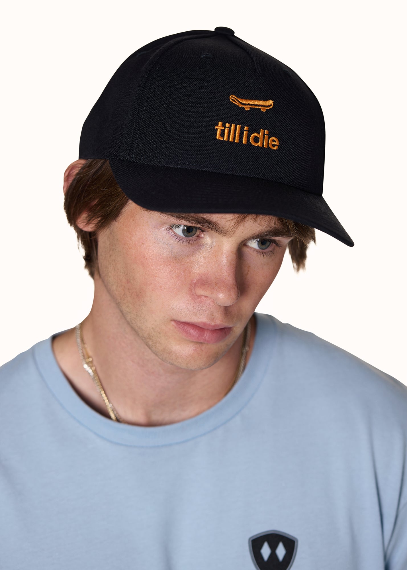 male model wearing a black snapback cap with an embroidered orange skateboard graphic with "till i die"