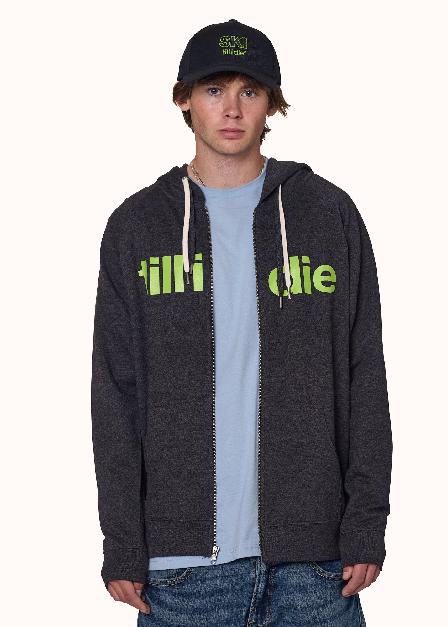 male model wearing a charcoal zip hoodie with lime green till i die written on the front, paired with a black ski till i die snapback cap and stone wash denim tee shirt