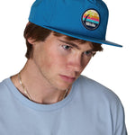 model wearing the marine blue sunset session surf hat, the hat has a pvc round patch with a palm tree and sunset graphic with the word "till i die"
