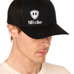 Front view of a male model wearing a black classic trucker hat, featuring a white embroidered Till I Die logo on the lower right front panel.