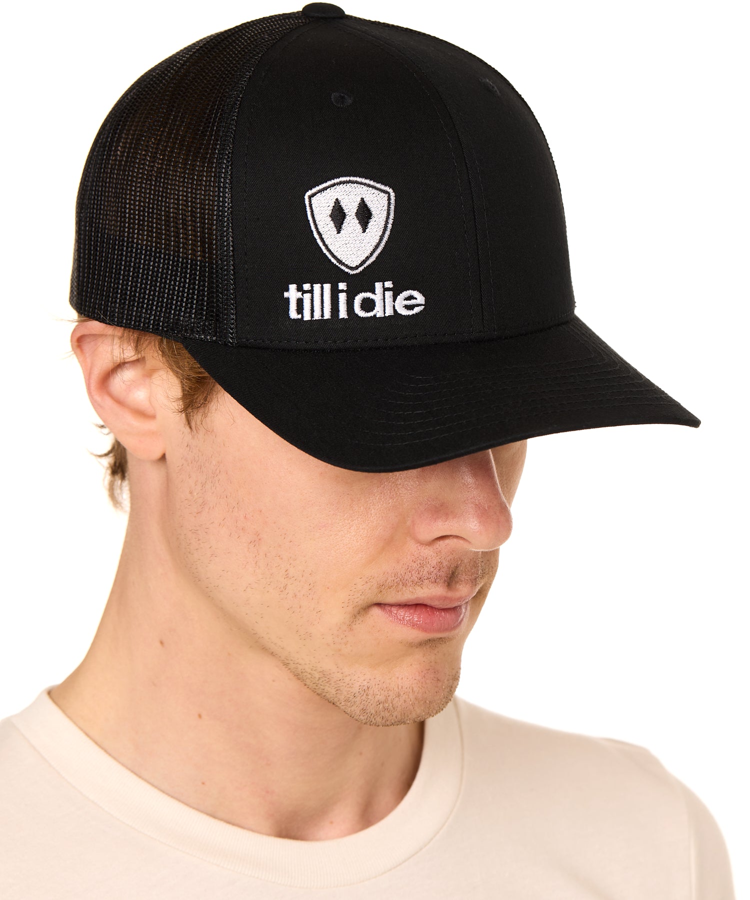Front view of a male model wearing a black classic trucker hat, featuring a white embroidered Till I Die logo on the lower right front panel.
