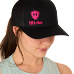 Front view of a female model wearing a black classic trucker hat, featuring a hot pink embroidered Till I Die logo on the lower right front panel.