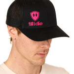 Front view of a male model wearing a black classic trucker hat, featuring a hot pink embroidered Till I Die logo on the lower right front panel.