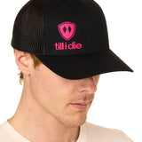 Front view of a male model wearing a black classic trucker hat, featuring a hot pink embroidered Till I Die logo on the lower right front panel.