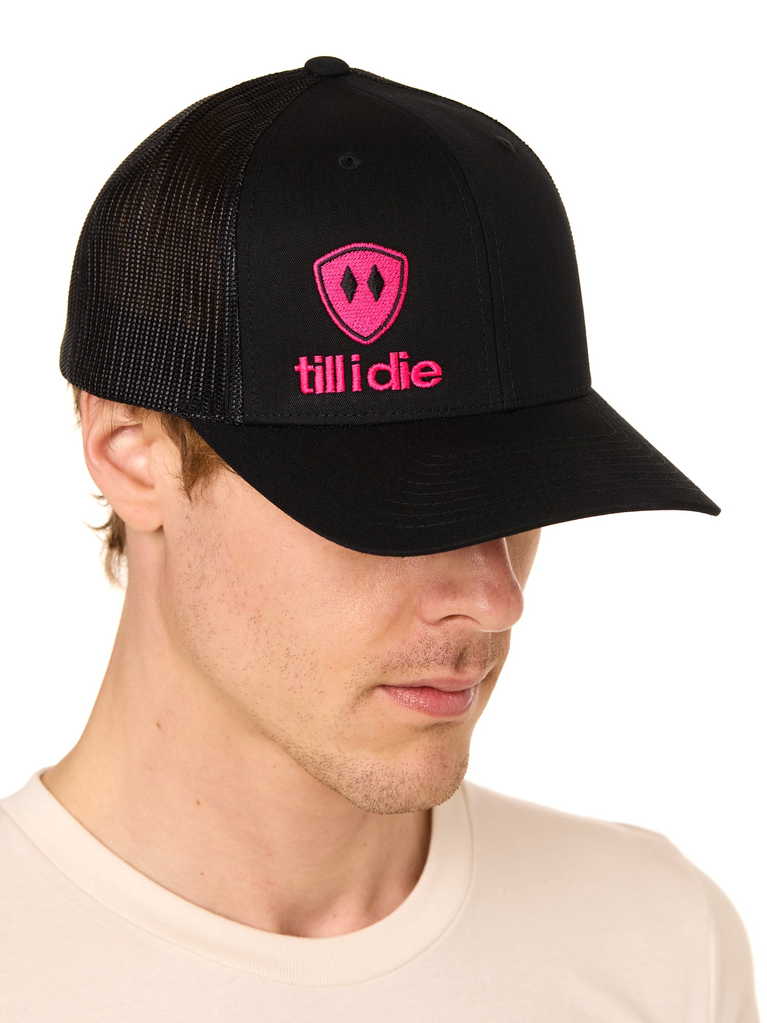 Front view of a male model wearing a black classic trucker hat, featuring a hot pink embroidered Till I Die logo on the lower right front panel.