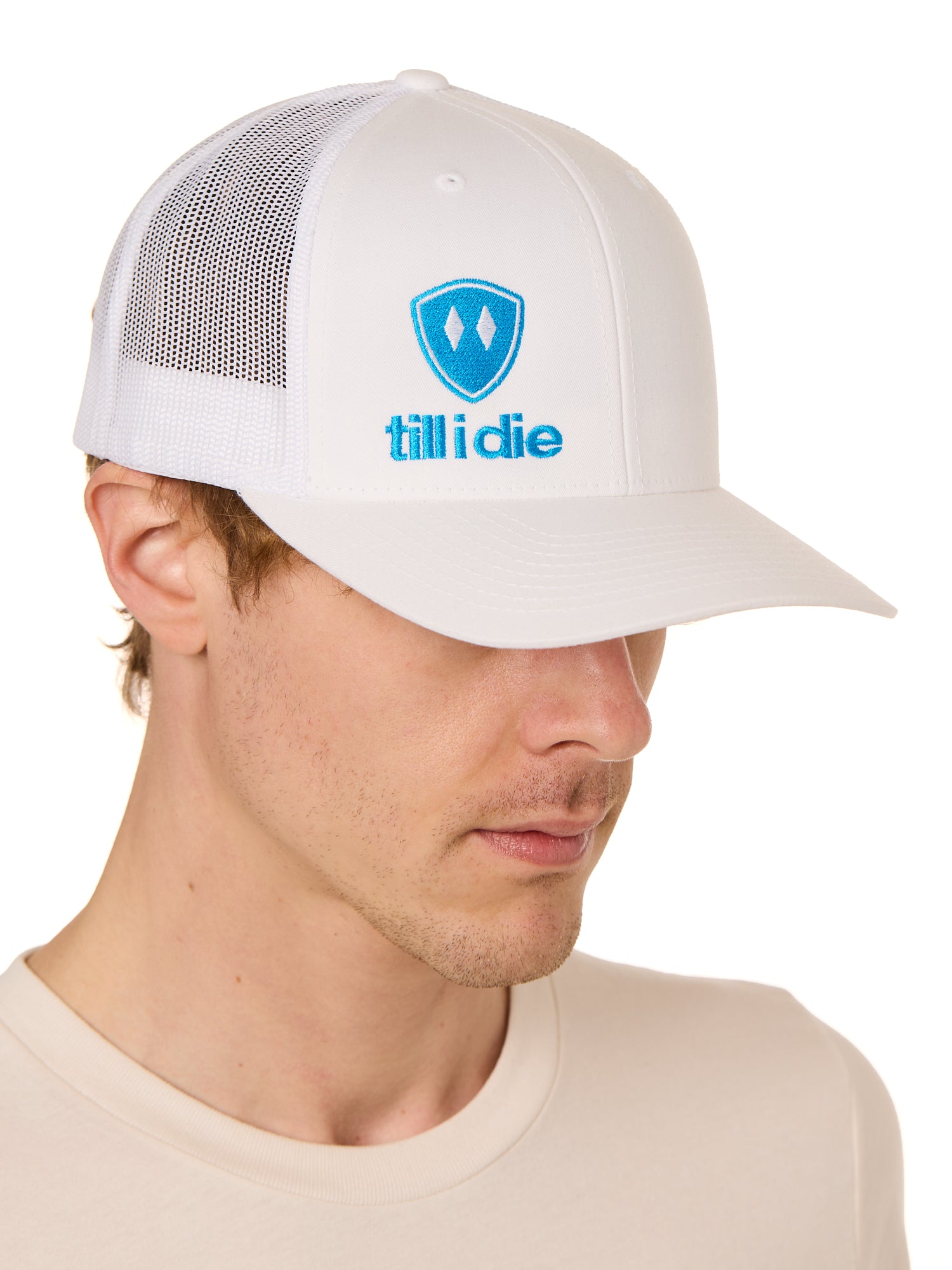 Front view of a male model wearing a white classic trucker hat, featuring a blue embroidered Till I Die logo on the lower right front panel.