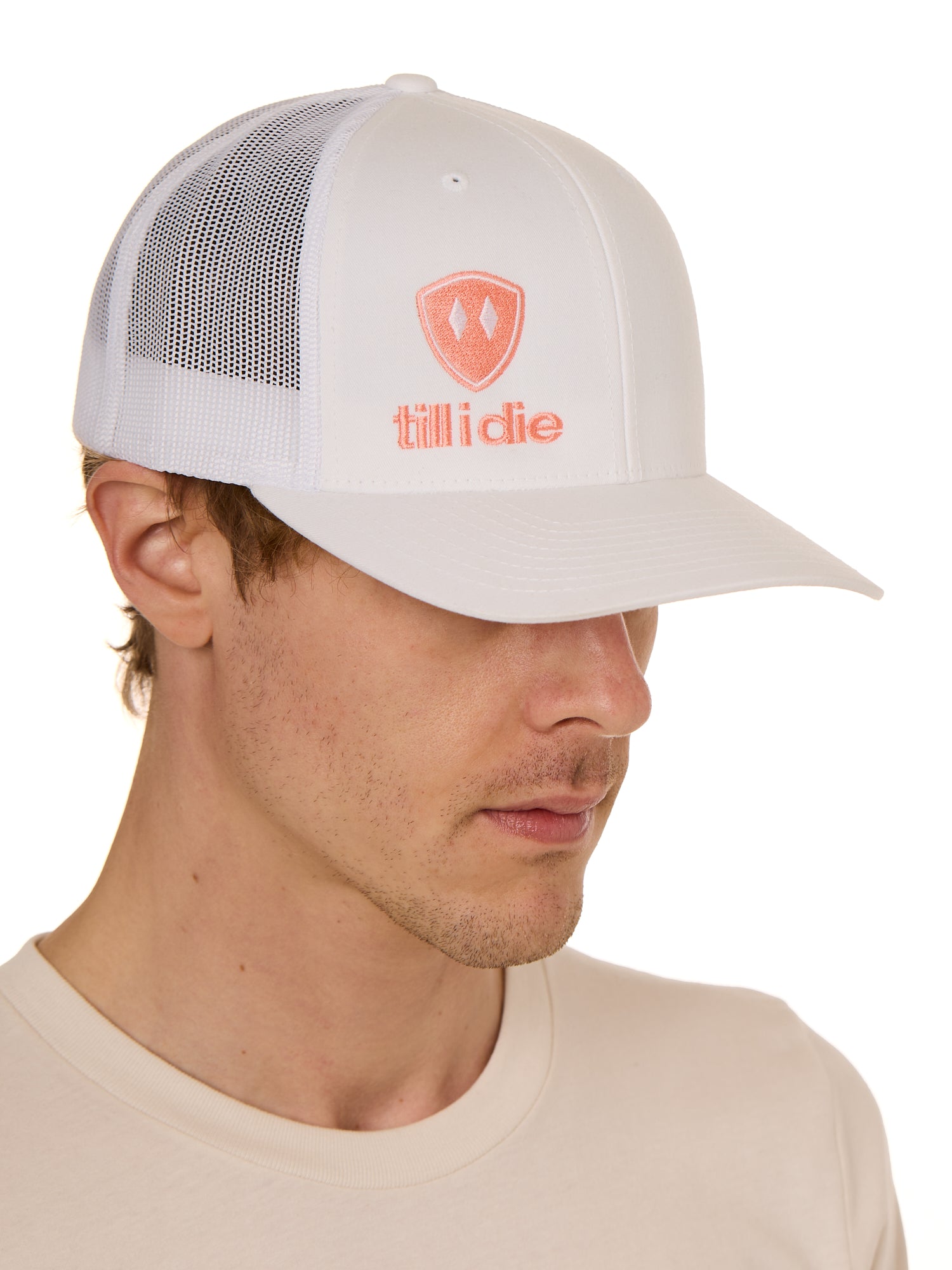 Front view of a male model wearing a white classic trucker hat, featuring a coral embroidered Till I Die logo on the lower right front panel.