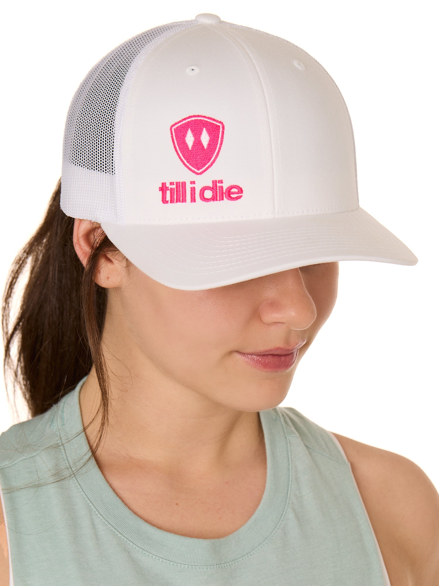 Front view of a female model wearing a white classic trucker hat, featuring a hot pink embroidered Till I Die logo on the lower right front panel.