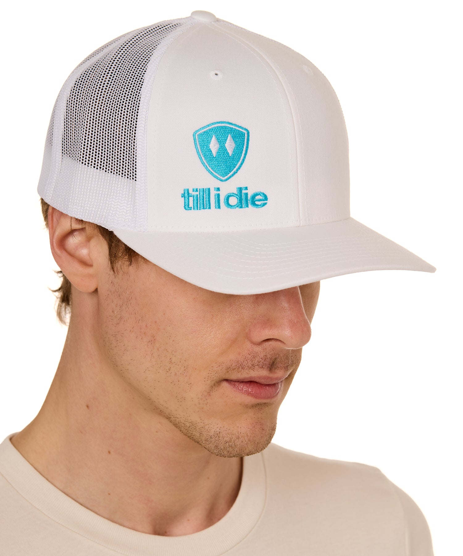 Front view of a male model wearing a white classic trucker hat, featuring a teal embroidered Till I Die logo on the lower right front panel.