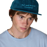 model wearing the corduroy surf hat in teal, has has light teal embroidery of a surf board and the words 'wet and wrecked'