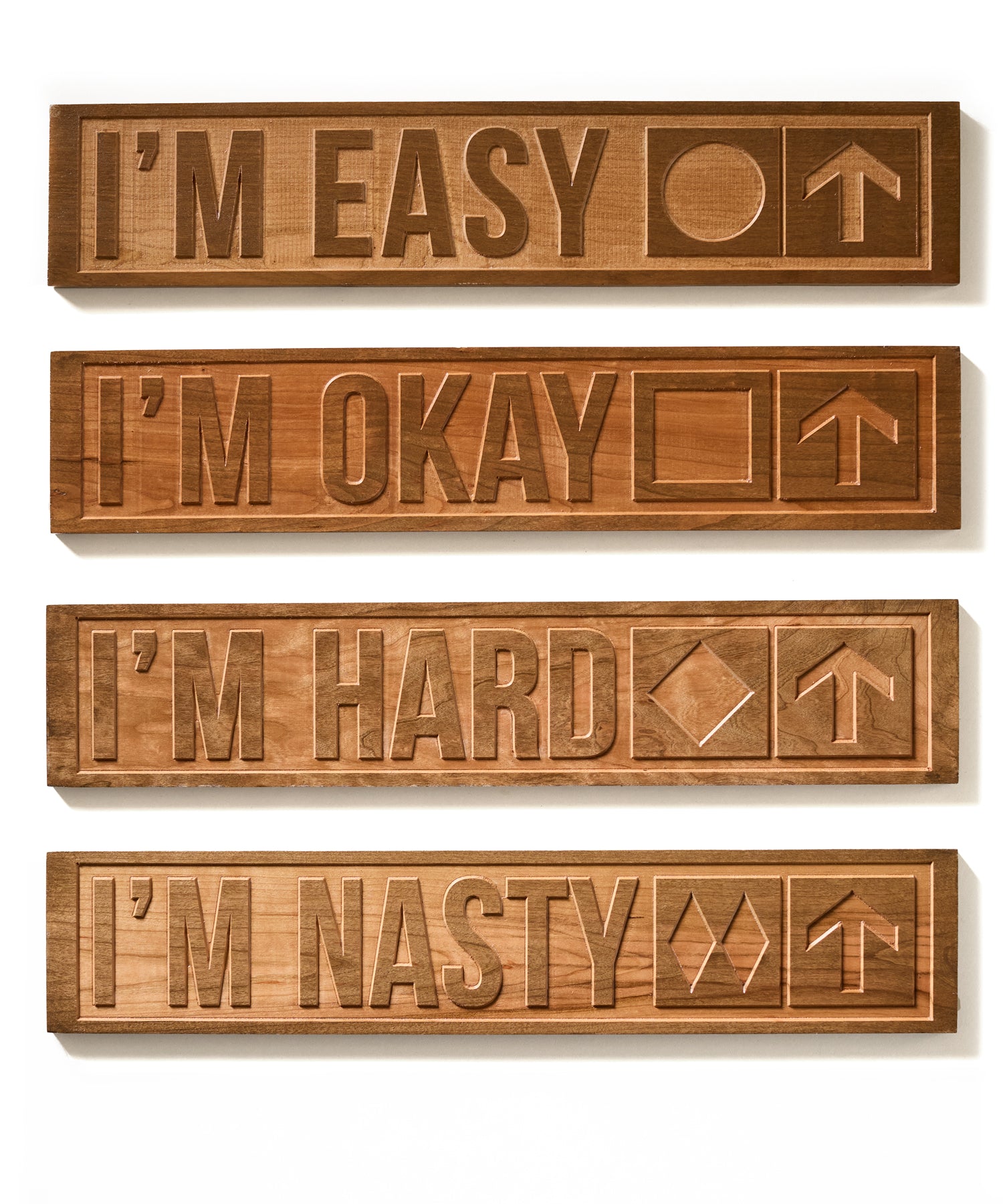 Collection of four cherry wood-carved ski trail signs displayed on a white background. Each sign features a different trail difficulty: “I’m Easy” with a green circle, “I’m Okay” with a blue square, “I’m Hard” with a black diamond, and “I’m Nasty” with double black diamonds—each paired with an upward-pointing arrow.