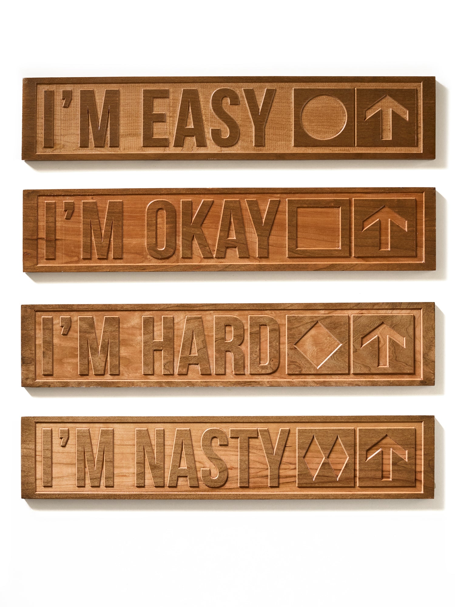 Collection of four cherry wood-carved ski trail signs displayed on a white background. Each sign features a different trail difficulty: “I’m Easy” with a green circle, “I’m Okay” with a blue square, “I’m Hard” with a black diamond, and “I’m Nasty” with double black diamonds—each paired with an upward-pointing arrow.