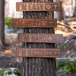 Wooded scene featuring four cherry wood-carved ski trail signs mounted on a tree. Each sign displays a different trail difficulty: “I’m Easy” with a green circle, “I’m Okay” with a blue square, “I’m Hard” with a single black diamond, and “I’m Nasty” with double black diamonds—all with arrows pointing upward.