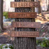 Wooded scene featuring four cherry wood-carved ski trail signs mounted on a tree. Each sign displays a different trail difficulty: “I’m Easy” with a green circle, “I’m Okay” with a blue square, “I’m Hard” with a single black diamond, and “I’m Nasty” with double black diamonds—all with arrows pointing upward.